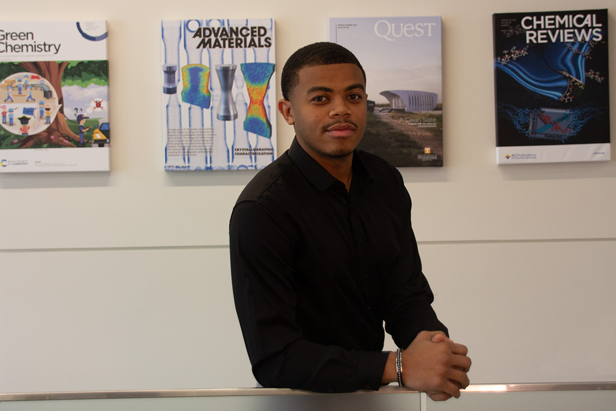 A young black man wearing a black dress shirt and a silver bracelet on his right wrist. He leans forward against a silver railing. Behind him is a light colored wall with canvas prints of journal covers. He looks at the camera with a pleasant neutral expression.