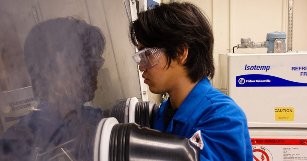 A young Asian man wearing a blue lab coat and clear safety glasses is using a glove box in a lab. A shadow of his image is reflected in the glove box window