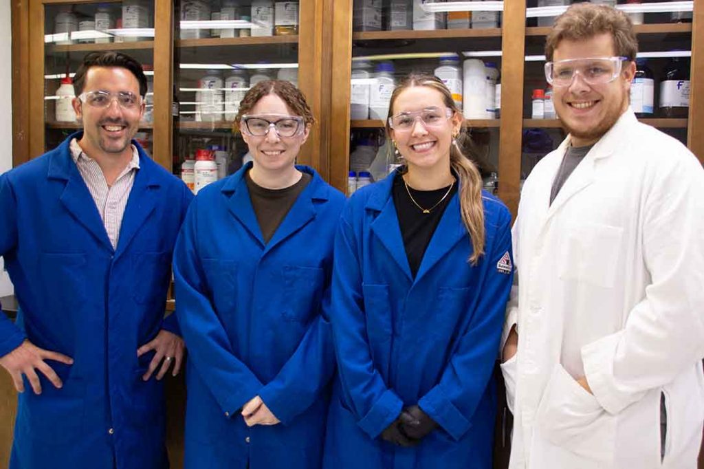 Dr. Baccile and graduate student Zack Hulsey stand with two young women in front of a case of lab supplies. They are wearing lab coats and safety glasses and are all smiling.