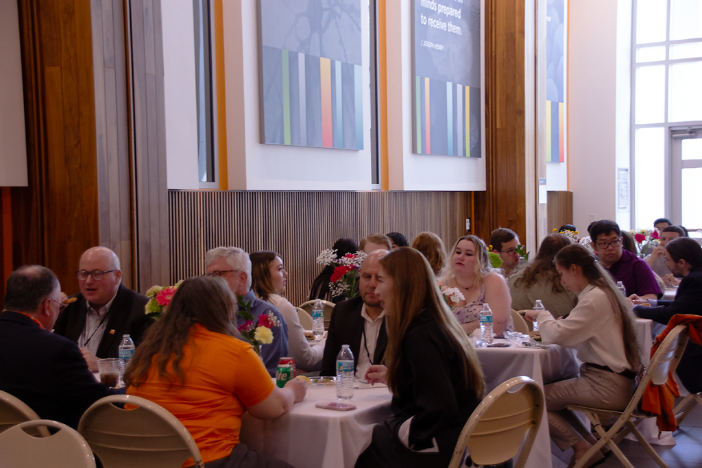 Symposium attendees sit at long tables in the Mossman Hall lobby having lunch.
