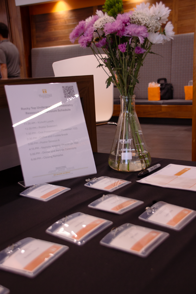 A table covered in a black tablecloth holds an upright copy of the symposium schedule, a glass scientific flask filles with purple and white flowers, and nametags for attendees.