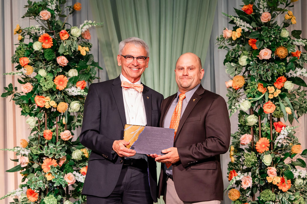 Professor Michael Best stands with Provost John Zomchick. They are holding the award and they are flanked by vertical floral arrangements containing multiple shades of orange, white, and green.