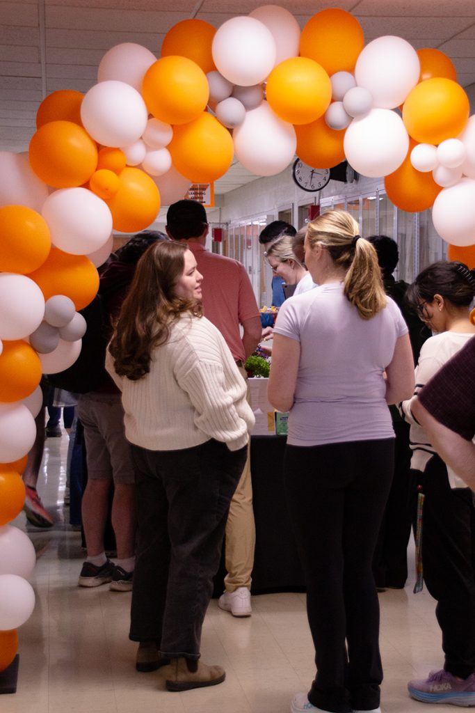 Graduate students stand in line for food beneath a balloon arch composed of orange, white, and gray balloons.