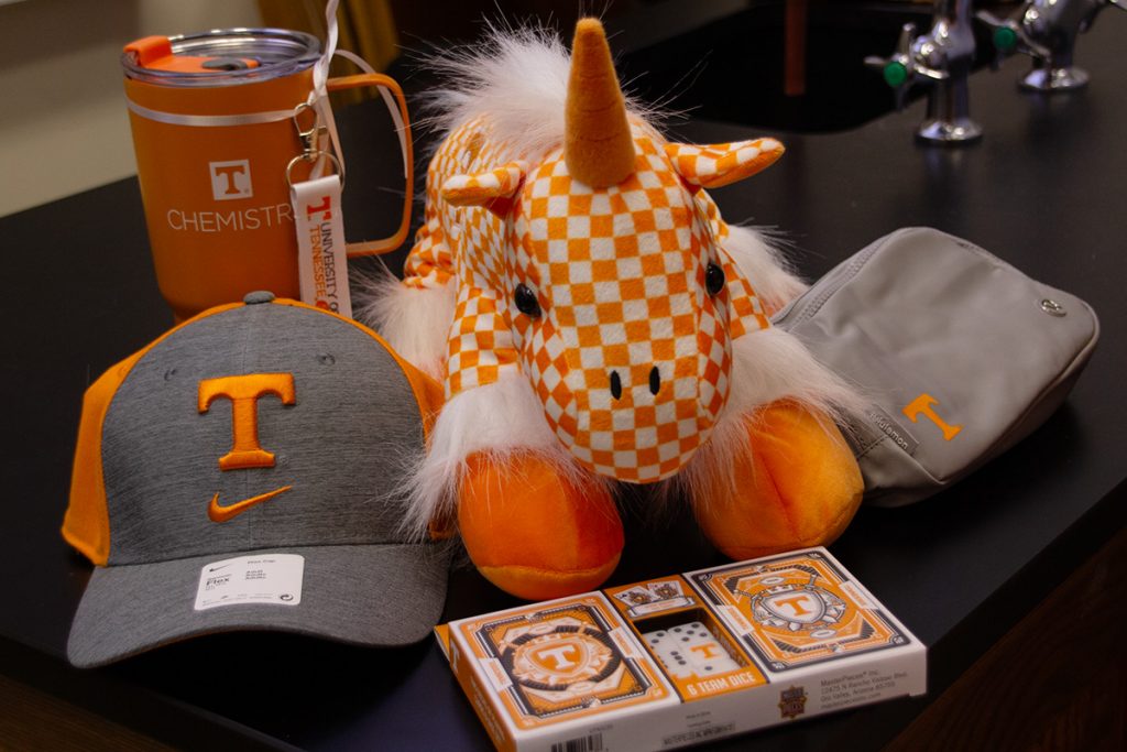 A group of raffle gifts sits on a dark table, including an orange Chemistry branded tumbler, a gray and orange UT Nike hat, a gray belt bag with a power T, a pack of UT branded cards and dice, and an orange and white checkered stuffed unicorn with an orange horn.