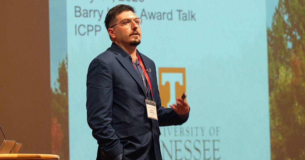 Dr. Brendon McNicholas stands in front of a large screen delivering the Barry Lever Young Investigator Award talk. He wears a navy blue suit and is looking out toward the audience.